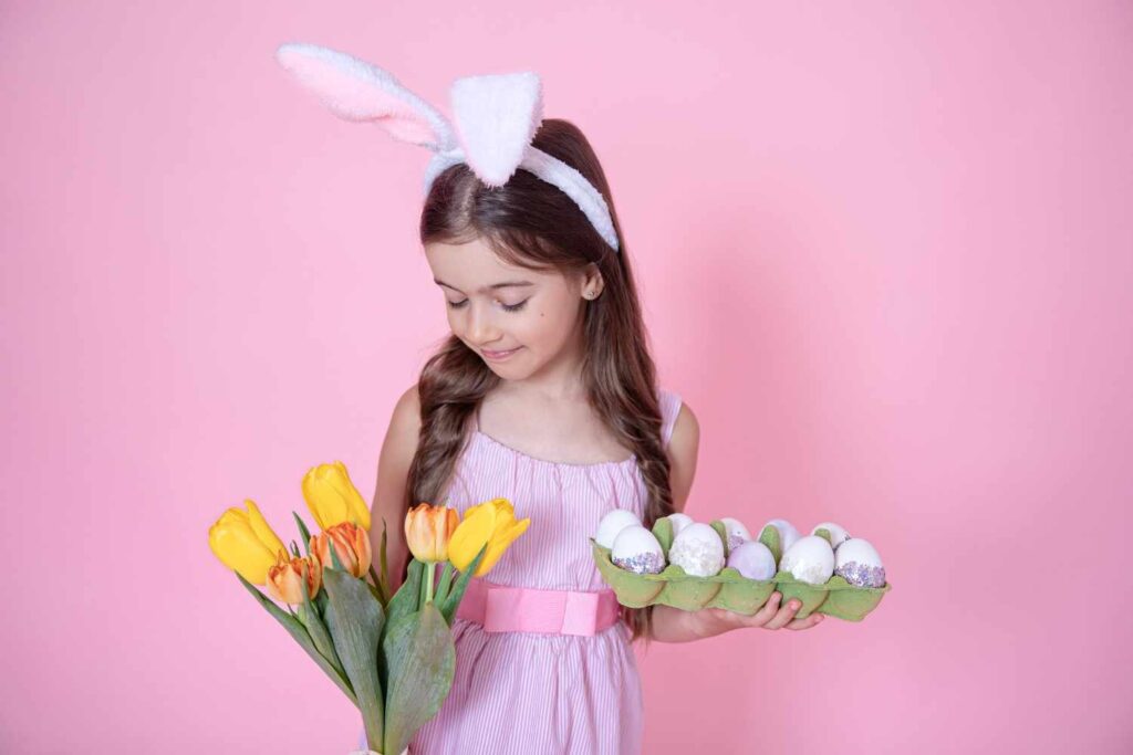 Girl wearing bunny ears holding Easter eggs and yellow tulips on pink background