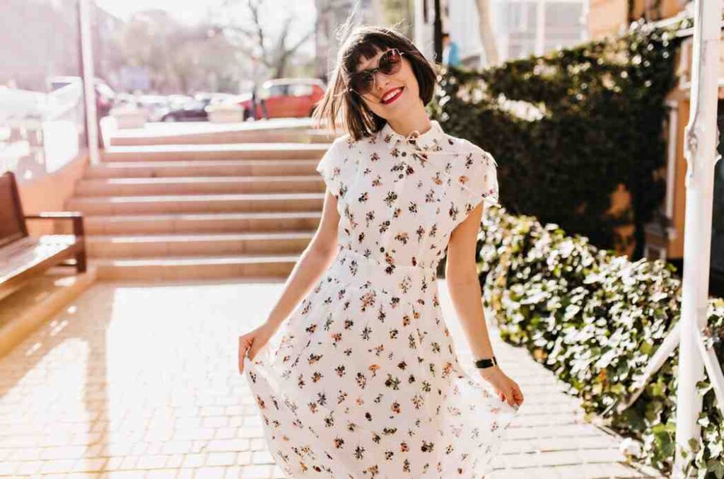 Woman smiling in white floral dress and sunglasses standing outside in sunlight
